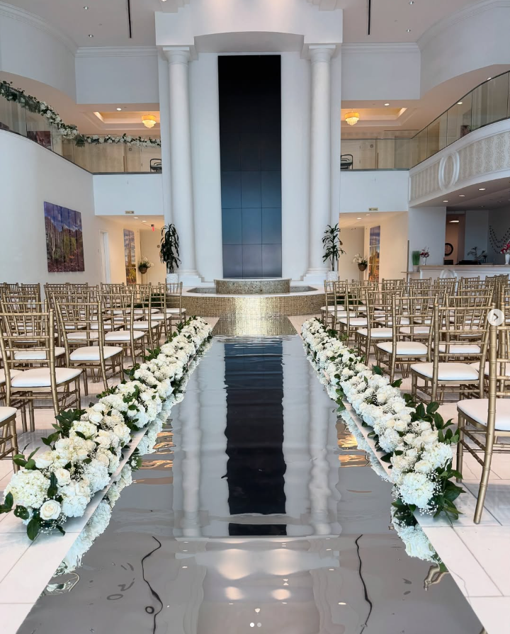 A wedding ceremony is taking place in a large room with a long aisle lined with chairs and flowers.