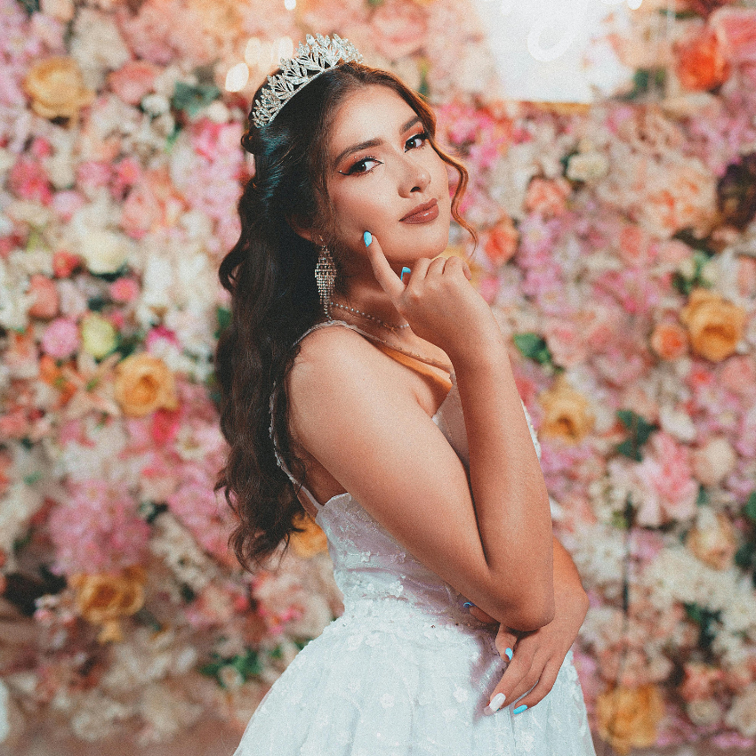 A woman in a white dress and tiara is standing in front of a wall of flowers.